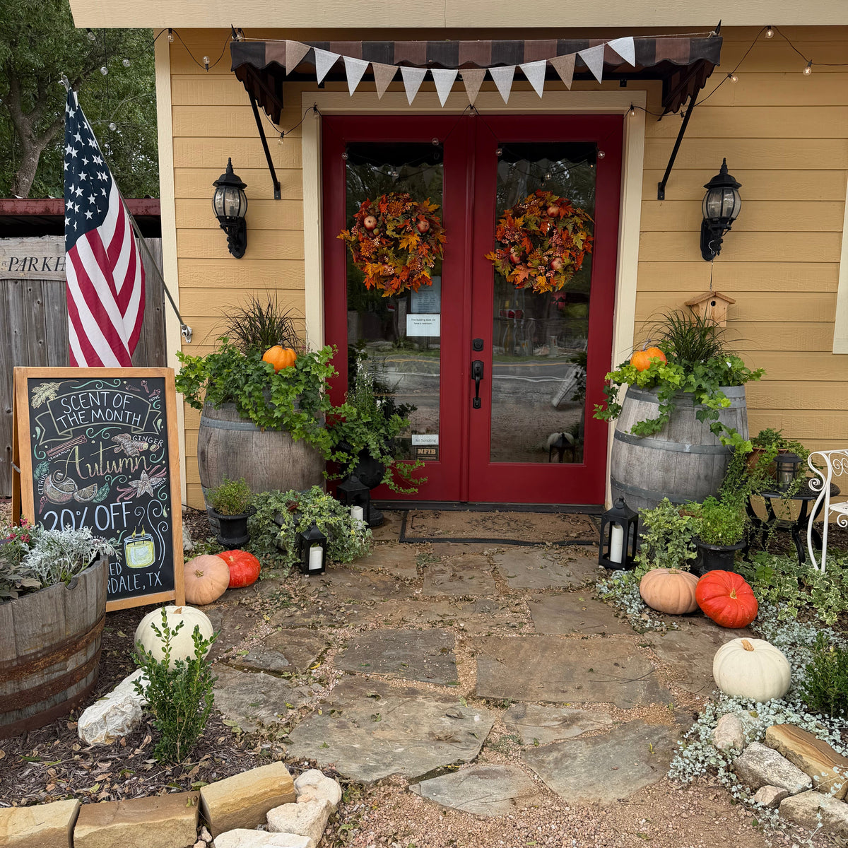 A styled fall porch scene in front of a yellow house with red double doors, decorated with autumn wreaths, pumpkins, plants, lanterns, a chalkboard sign, and an American flag.